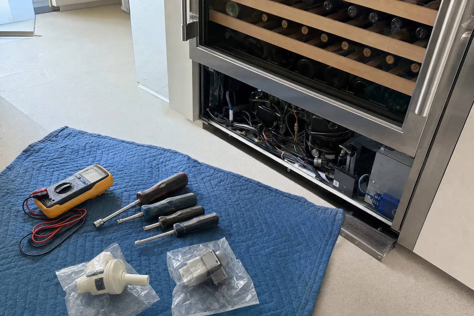 Undercounter wine cooler full of bottles with its lower grille removed, compressor exposed and tools laid out on a blanket