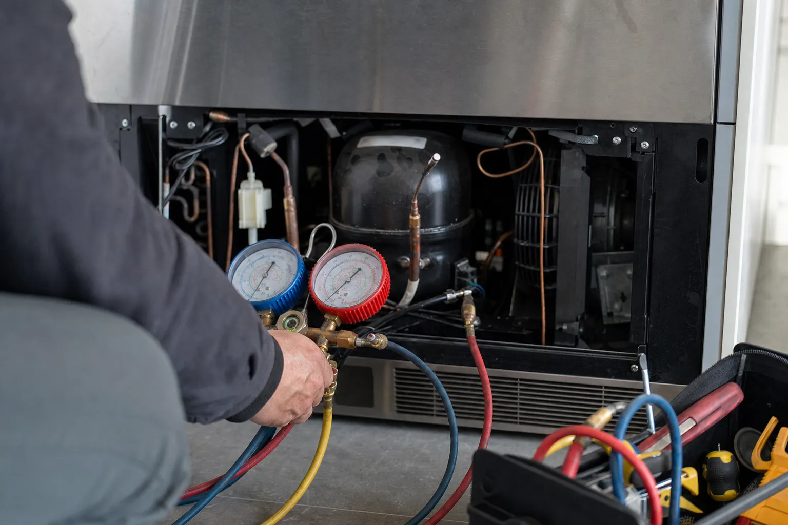 Refrigeration manifold gauges held at the open base of a stainless unit, hoses running to the exposed compressor