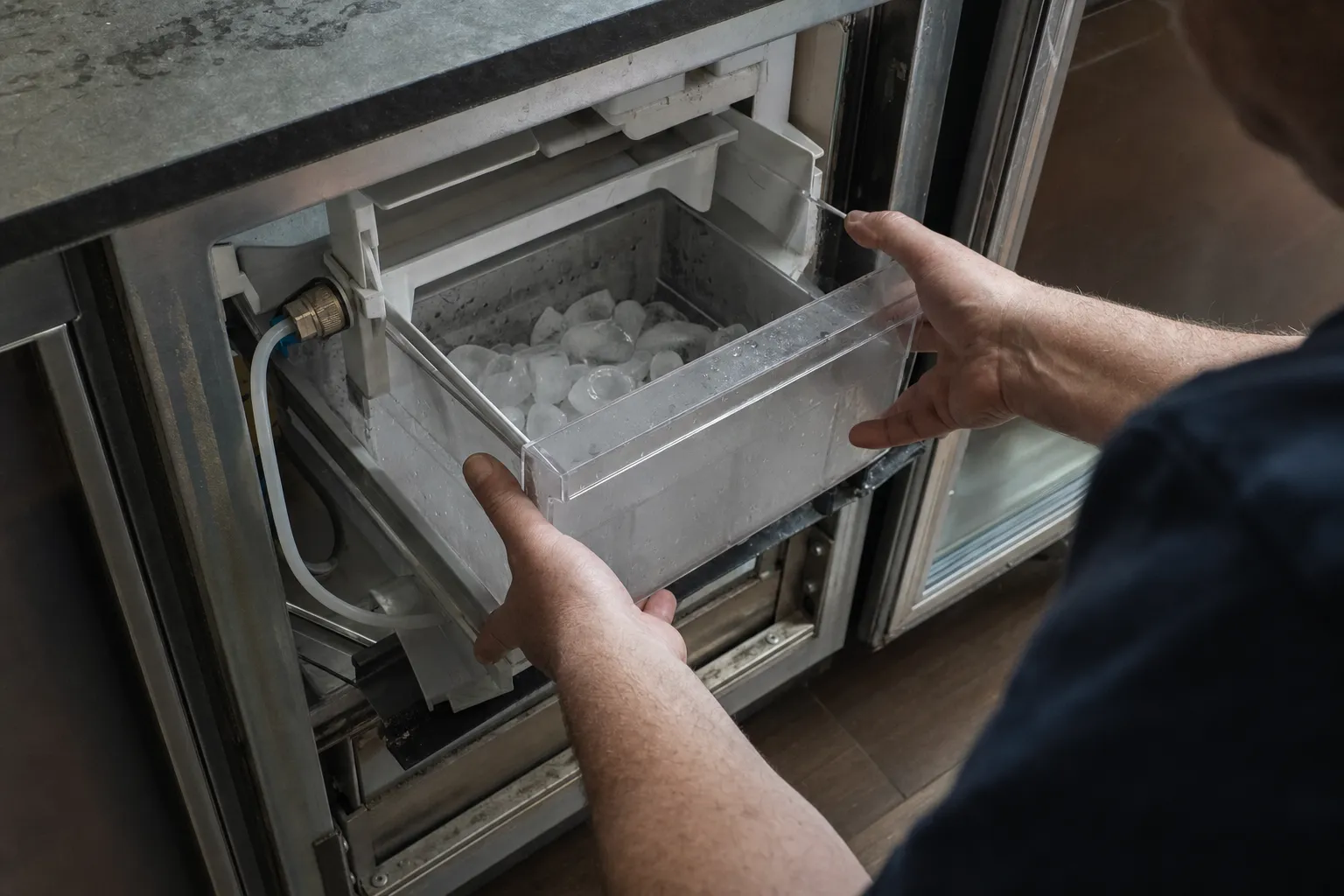 Clear ice bin of cubes drawn out of an undercounter unit, a water tube and brass fitting at the left of the opening